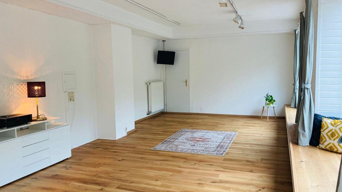 Ein helles, gemütliches Zimmer mit Holzfußboden und großen Fenstern. Es gibt eine kleine Pflanze und eine dekorative Sitzecke. | © Kathrin Leitner