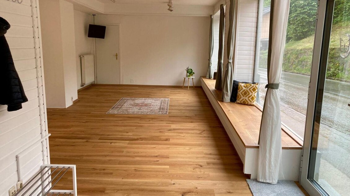 Ein heller, leerer Raum mit Holzfußboden und großen Fenstern. Ein Teppich und einige Sitzmöglichkeiten vervollständigen die gemütliche Atmosphäre. | © Kathrin Leitner