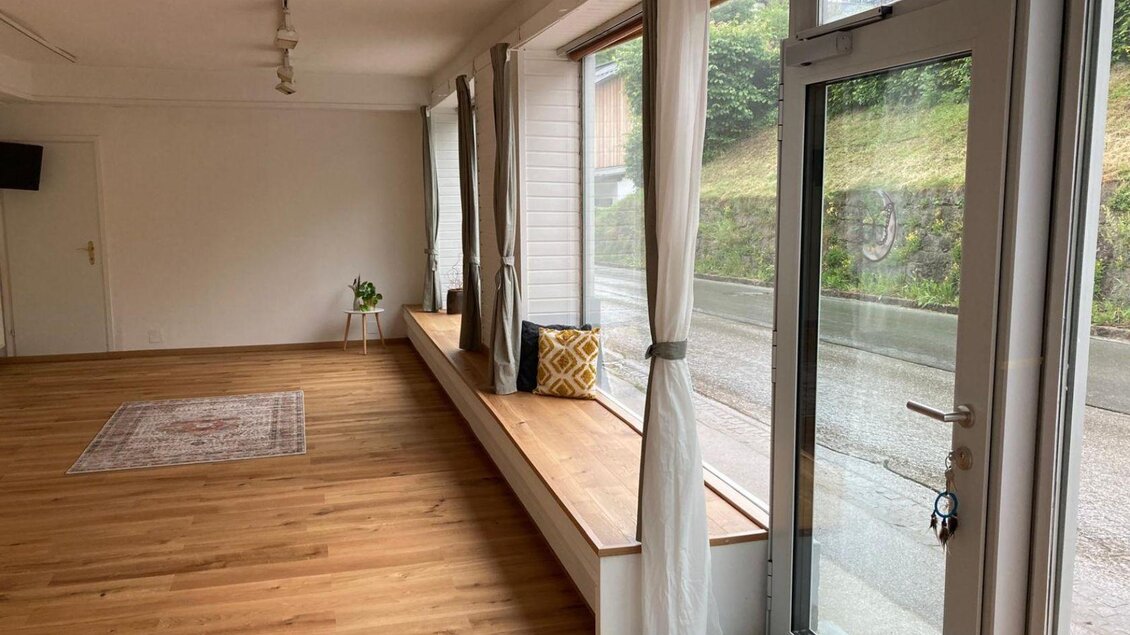 Ein heller Raum mit Holzboden und großen Fenstern. Auf der Fensterbank steht ein Kissen und eine kleine Pflanze. | © Kathrin Leitner