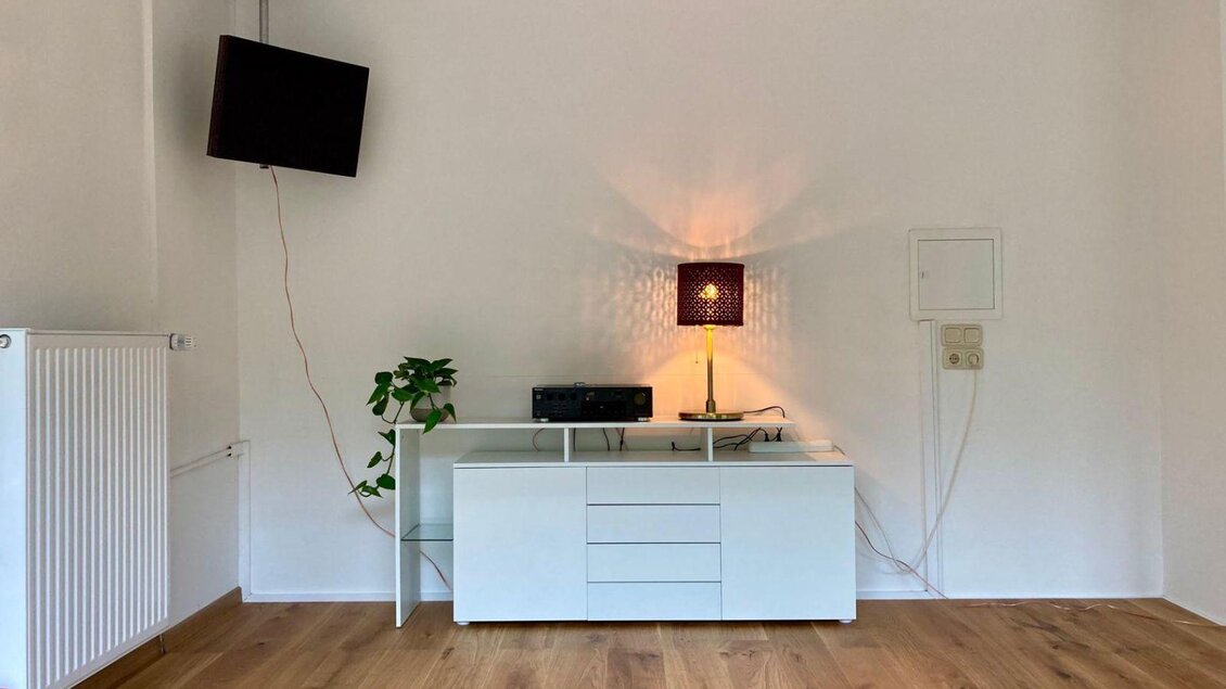 Ein heller Raum mit einem weißen TV-Rack und einem Fernseher. Auf dem Rack steht eine Lampe und eine kleine Pflanze. | © Kathrin Leitner
