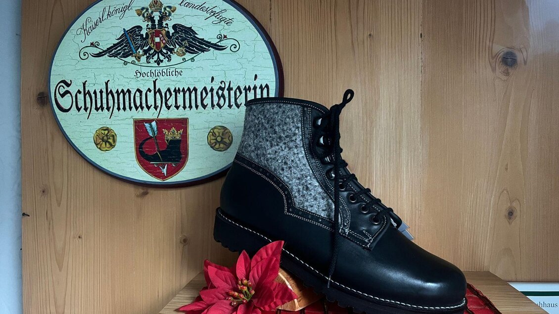 Ein handgefertigter Schuh aus Leder und Filz steht auf einem Tisch. Im Hintergrund ist ein Schild mit dem Schriftzug "Schuhmachermeisterin" zu sehen. | © TVB Ausseerland- Salzkammergut_Nathalie Schröttenhamer