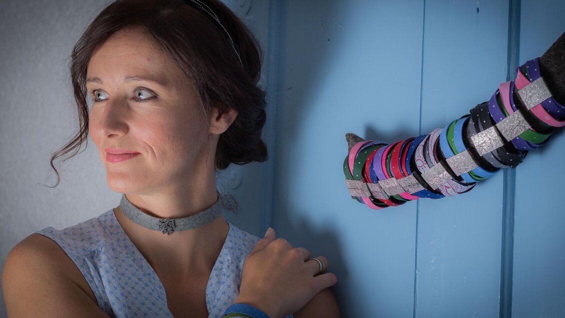 Eine Frau mit braunen Haaren und einem hellen Kleid steht vor einer blauen Wand. An der Wand hängen bunte Armbänder. | © Christina Neuper