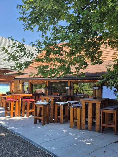 A cozy outdoor area with wooden furniture and plenty of greenery. In the background, there is a building with windows and a clear blue sky. | © TV Hochsteiermark