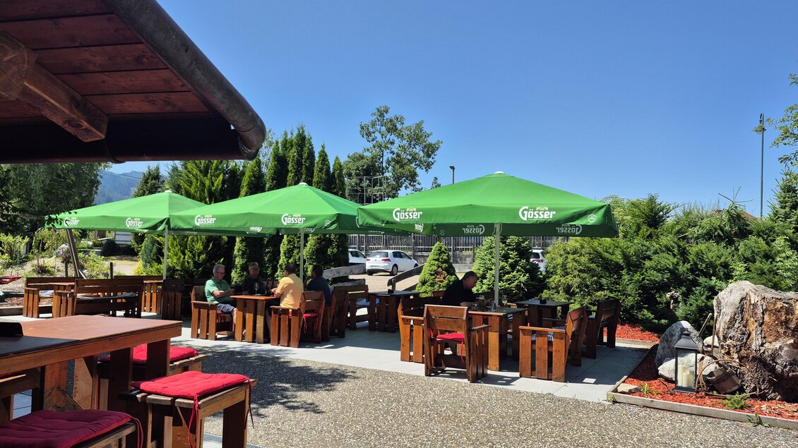 Ein gemütlicher Außenbereich mit Holztischen und grünen Markisen. Im Hintergrund sind Bäume und Sträucher zu sehen. | © TV Hochsteiermark