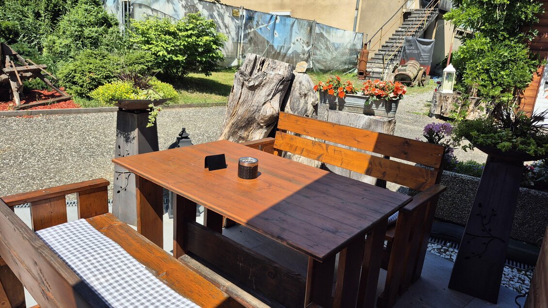 Ein gemütlicher Sitzbereich aus Holz im Freien mit einem Tisch und Bänken. Im Hintergrund sieht man eine grüne Gartenlandschaft und eine Wand mit Pflanzen. | © TV Hochsteiermark