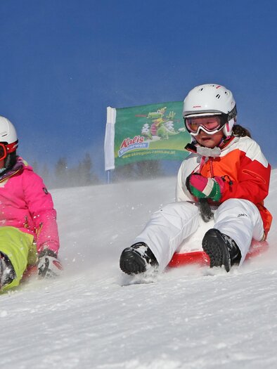 Two girls are happily sliding down the snowy slope. They are wearing helmets and colorful winter clothing. | © Tritscherhof