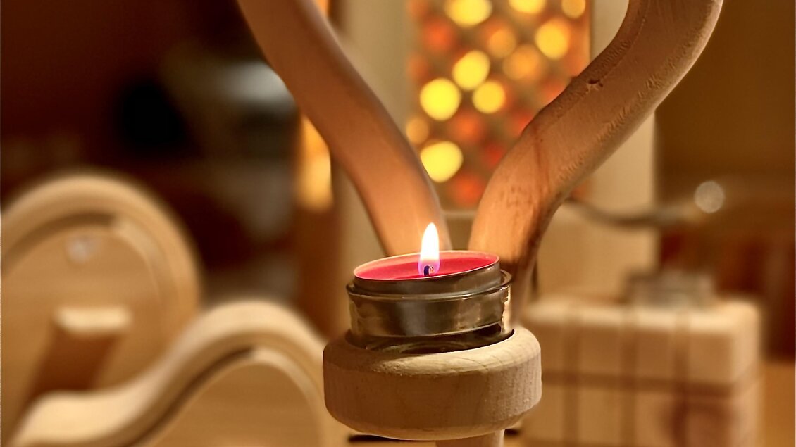 Eine Holzkerzenhalter in Herzform mit einer roten Teelichtkerze. Im Hintergrund sind weitere dekorative Holzobjekte zu sehen. | © Zirben Toni