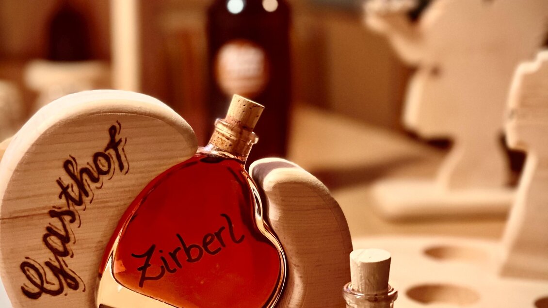 Zwei herzförmige Flaschen mit einem rotbraunen Getränk stehen auf einem Tisch. Im Hintergrund sind handgemachte Holzfiguren und eine brennende Kerze sichtbar. | © Zirben Toni