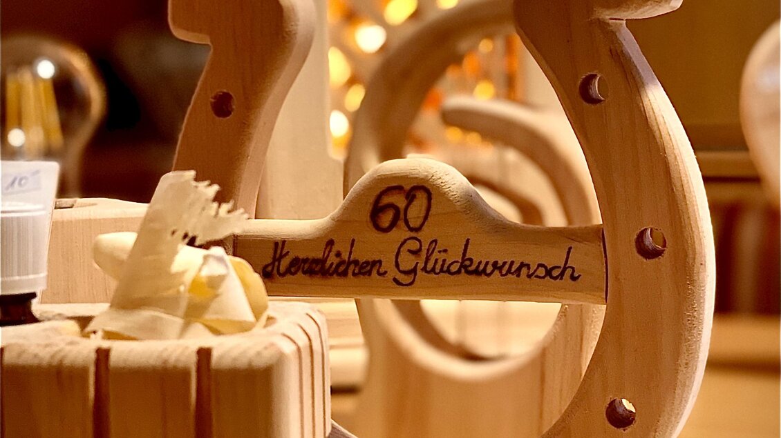 Ein hölzernes Glückssymbol in Form eines Hufeisens mit der Aufschrift „60. Herzlichen Glückwunsch“. Im Hintergrund sind dekorative Elemente sichtbar, die eine festliche Atmosphäre schaffen. | © Zirben Toni