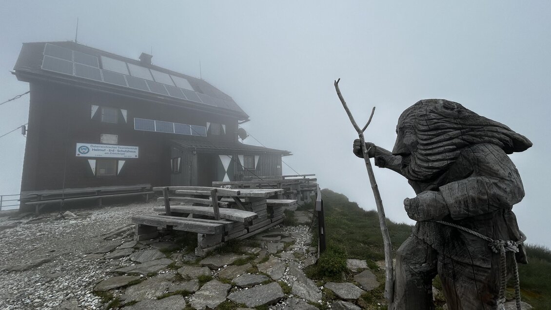 Eine Berghütte umgeben von Nebel, mit einem Holzschnitzel des Bergführers im Vordergrund. Der Weg ist mit Steinen bedeckt und bietet eine mystische Atmosphäre. | © Tourismusverband Murau