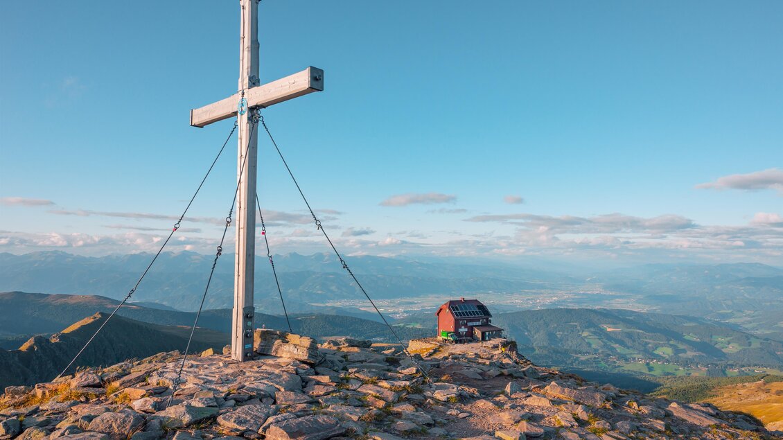Ein Gipfel mit einem großen Kreuz und einer kleinen Hütte. Im Hintergrund erstrecken sich die Berge und der Himmel ist klar und blau. | © TVB Murau