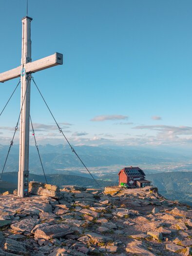A peak with a large cross and a small hut. In the background, the mountains extend and the sky is clear and blue. | © TVB Murau
