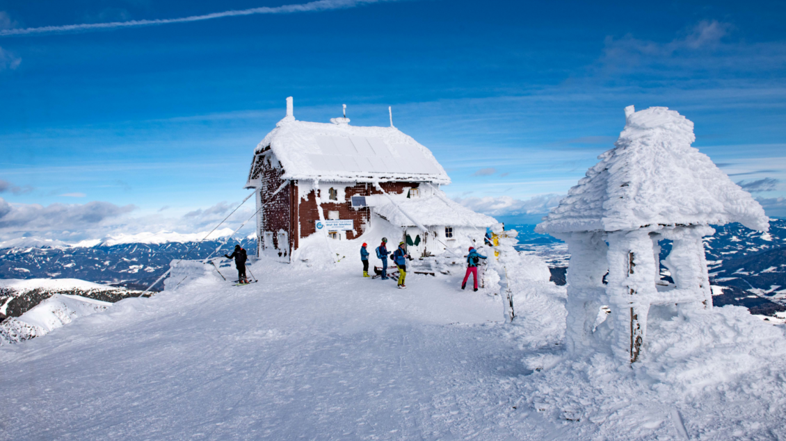 Eine verschneite Berglandschaft mit einem gemütlichen Holzhaus und mehreren Personen, die Ski fahren. Der Himmel ist klar und blau, und die Berge sind im Hintergrund sichtbar. | © Anita Fössl