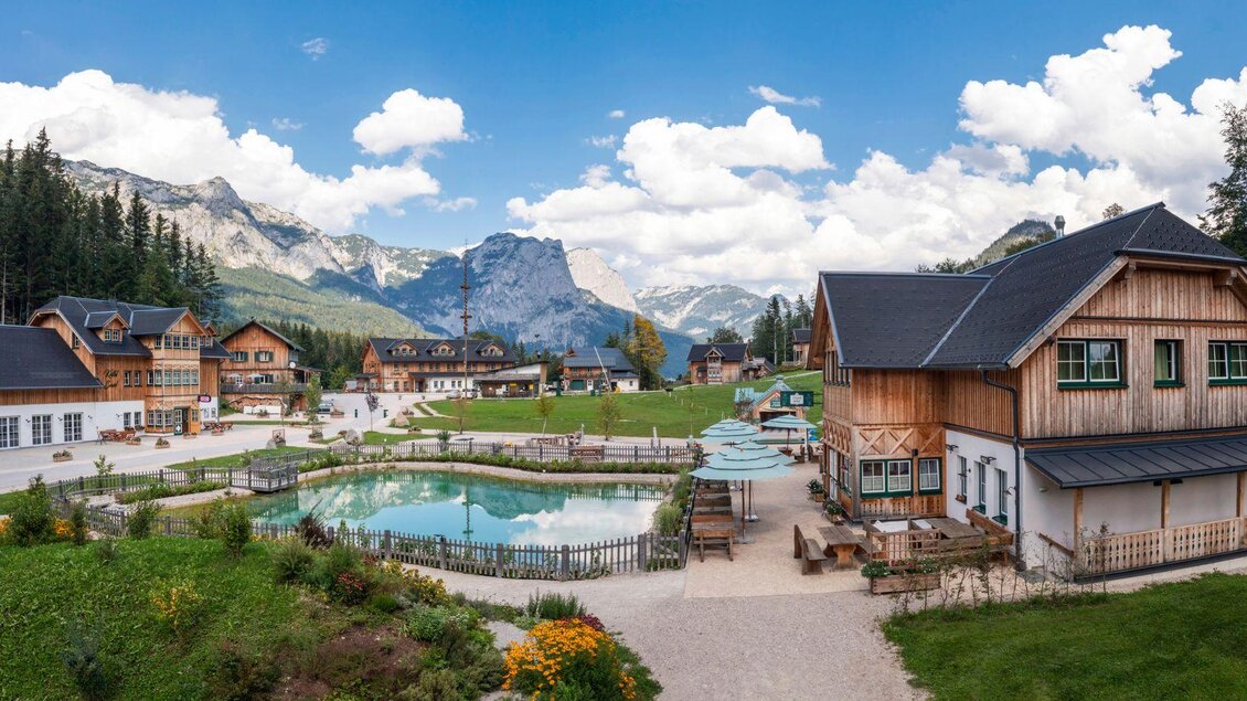 Eine malerische Landschaft mit traditionellen Holzhäusern und einem klaren Schwimmteich. Im Hintergrund erheben sich majestätische Berge unter einem blauen Himmel mit weißen Wolken. | © www.zloam.at