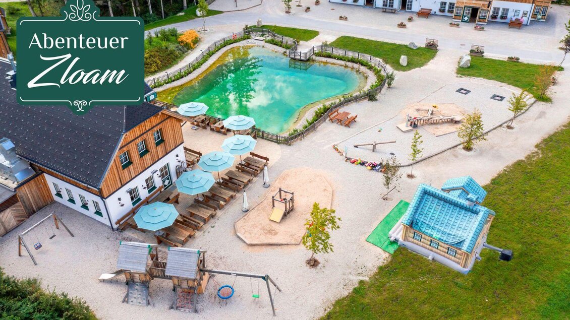 Ein großer Außenbereich mit einem grünen Teich, Spielplätzen und gemütlichen Sitzgelegenheiten. Im Hintergrund sind Holzgebäude und eine entspannende Landschaft zu sehen. | © www.zloam.at