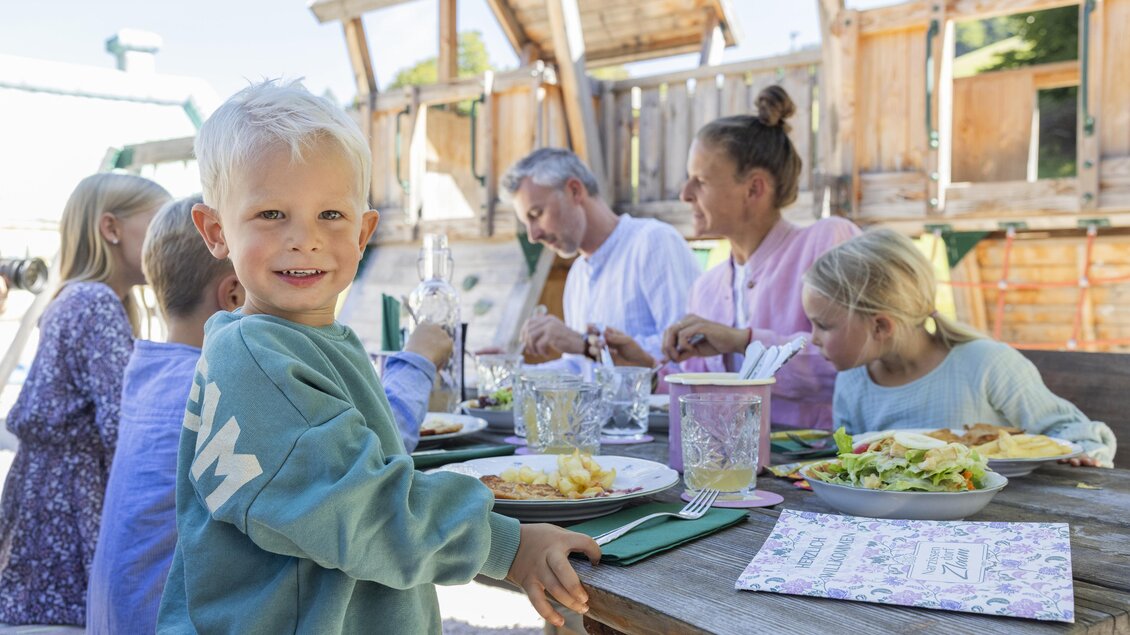 Eine fröhliche Familie isst gemeinsam im Freien. Ein Junge mit blonden Haaren lächelt am Tisch, während andere Familienmitglieder essen. | © www.zloam.at
