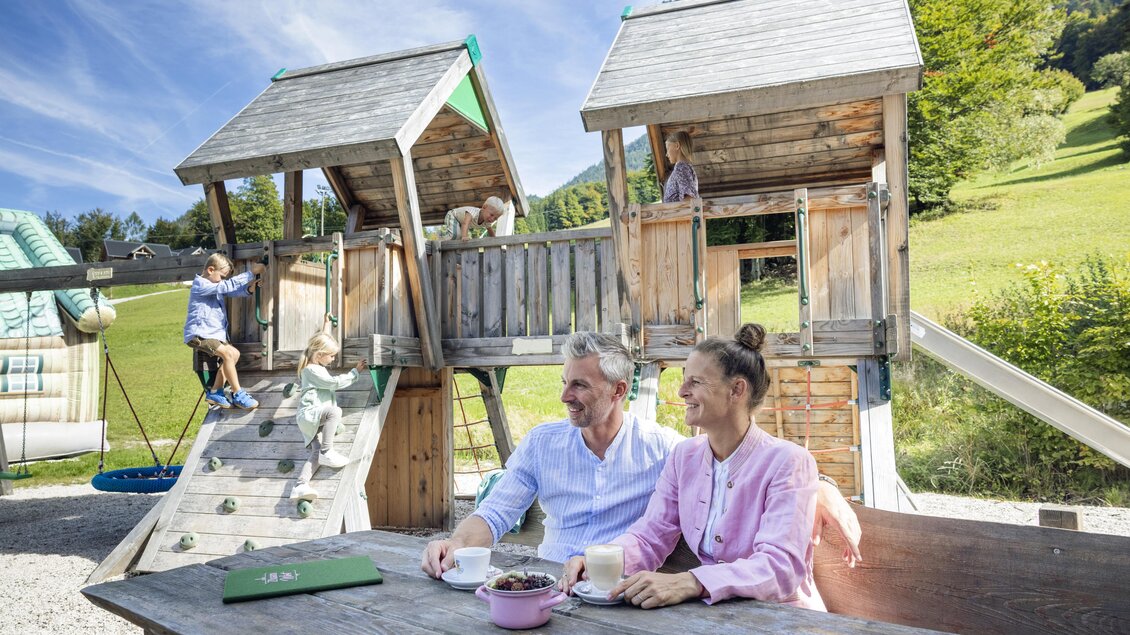 Ein Spielplatz mit zwei Holzspielhäusern und einer Rutsche. Im Vordergrund sitzen ein Paar mit Getränken und genießen die Aussicht auf die spielenden Kinder. | © www.zloam.at