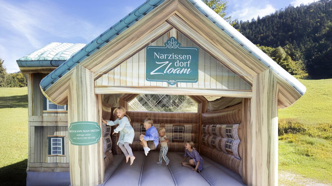 Ein aufblasbares Spielhaus mit dem Namen "Narzissen Dorf Zloam". Kinder spielen und hüpfen darin auf einer fröhlichen Wiese. | © www.zloam.at