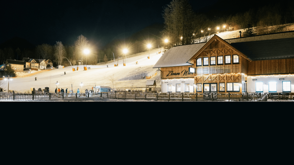 Eine verschneite Piste mit Beleuchtung bei Nacht. Im Vordergrund ist ein gemütliches Holzgebäude zu sehen.