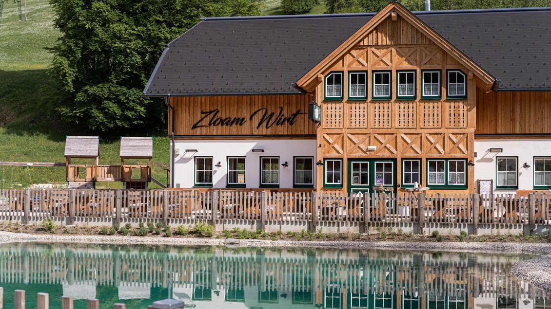 Ein rustikales Holzhaus mit mehreren Fenstern und einem großen Schriftzug ist am Ufer eines klaren Teichs zu sehen. Umgeben von grünen Bäumen und einer gemütlichen Atmosphäre. | © Zloam Wirt, www.zloamwirt.at