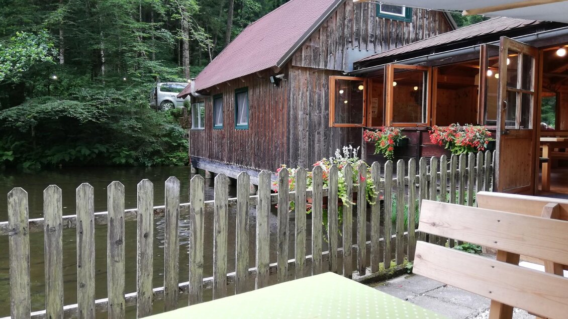 Ein rustikales Restaurant am Ufer eines Teiches, umgeben von Bäumen. Die gemütliche Terrasse mit vorbeigemalten Blumenkästen lädt zum Verweilen ein.
