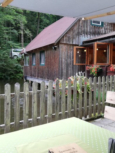 A rustic restaurant by the edge of a pond, surrounded by trees. The cozy terrace with painted flower boxes invites you to linger.