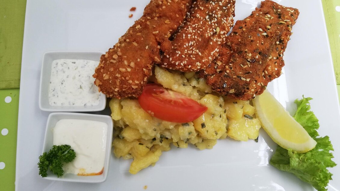 Ein Teller mit gebratenem Fischfilet, serviert mit Kartoffelsalat und frischen Beilagen. Dazu gibt es verschiedene Dips und eine Zitronenscheibe.