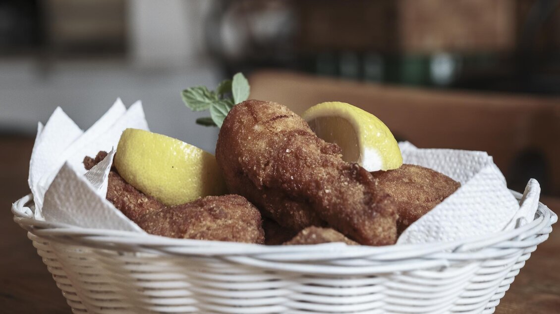 Ein Korb mit knusprigen Hühnchenteilen, die mit Zitronenscheiben serviert werden. Das Gericht ist auf einem Holzuntergrund platziert. | © Anton Muchitsch