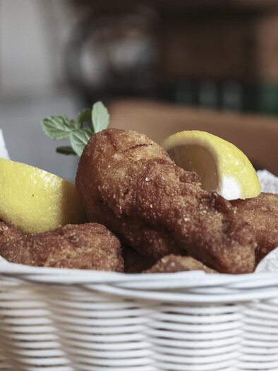 Ein Korb mit knusprigen Hühnchenteilen, die mit Zitronenscheiben serviert werden. Das Gericht ist auf einem Holzuntergrund platziert. | © Anton Muchitsch