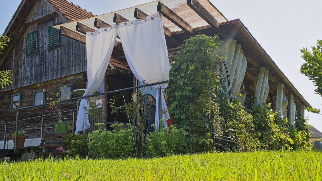 Ein gemütliches Holzhaus mit großzügiger Terrasse und Pflanzen. Weisse Vorhänge wehen sanft im Wind, umgeben von grünem Gras. | © Anton Muchitsch