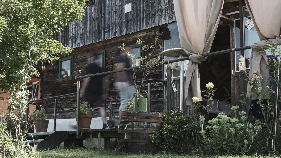 Ein rustikales Holzhaus mit Pflanzen und einem Eingang. Eine Person bewegt sich schnell in der Nähe des Hauses. | © Anton Muchitsch