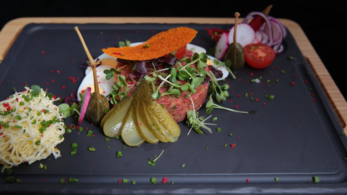 Ein kunstvoll angerichteter Teller mit einem Pattie, eingelegten Gurken, frischem Gemüse und einer knusprigen Dekoration. Daneben befinden sich Kräuter und eine Portion Käse. | © Zur Schmied'n - Kai Hasewend