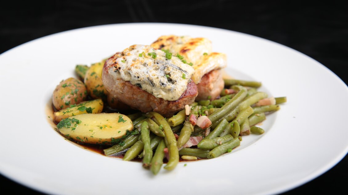 Ein leckerer Teller mit gegrilltem Fleisch, überbacken mit Käse, serviert mit grünen Bohnen und Kartoffeln. Ein appetitliches Gericht auf einem weißen Teller. | © Kai Hasewend
