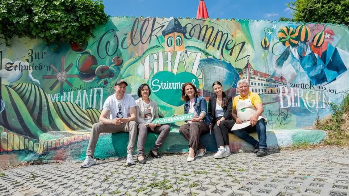 Gruppe vor farbigem Willkommens-Wandbild mit Graz- und Steiermark-Motiven. | © Zur Schmied'n