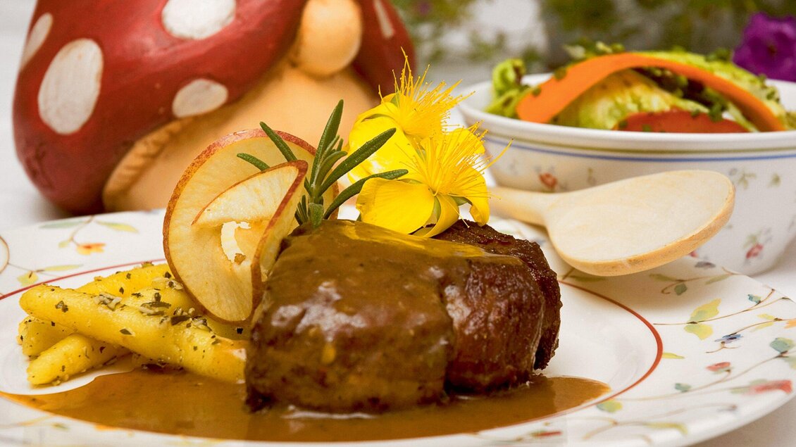 Ein köstliches Gericht mit zartem Fleisch, serviert mit einer Sauce und Beilagen aus Kartoffeln. Im Hintergrund sieht man einen bunten Salat und eine verspielte Pilzfigur. | © Steirerstub'n