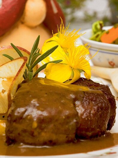 A delicious dish with tender meat, served with a sauce and side dishes of potatoes. In the background, you can see a colorful salad and a playful mushroom figure. | © Steirerstub'n