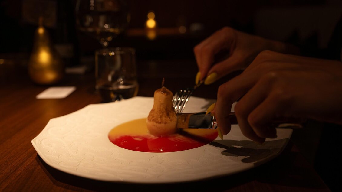 Hand schneidet pochierte Birne auf Dessertteller mit roter und gelber Sauce | © 5komma5sinne - Rene Strasser Fotografie