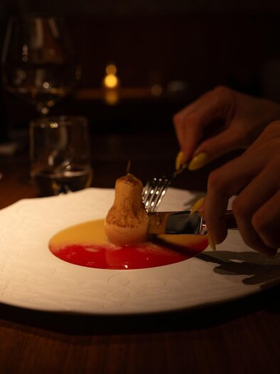 Hand schneidet pochierte Birne auf Dessertteller mit roter und gelber Sauce | © 5komma5sinne - Rene Strasser Fotografie