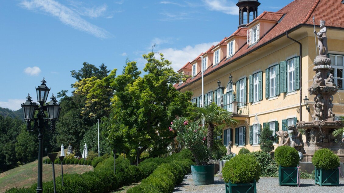 Ein historisches Gebäude mit grünen Fensterläden und einem roten Dach, umgeben von gepflegten Gärten. Im Vordergrund stehen elegante Lampen und kunstvolle Pflanzen. | © Aiola - Ladies & Lord