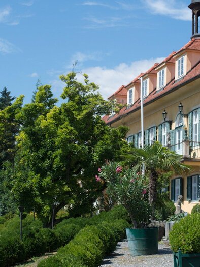 A historic building with green shutters and a red roof, surrounded by well-kept gardens. In the foreground are elegant lamps and artistic plants. | © Aiola - Ladies & Lord