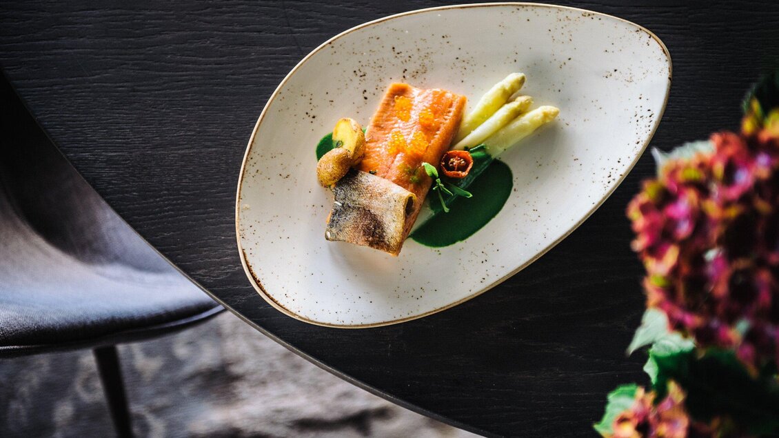 Ein kunstvoll angerichteter Teller mit gebratenem Fisch, Spargel und einer grünen Sauce. Dazu sind einige dekorative Gemüse und Kräuter platziert. | © Aiola - Joel Kernasenko