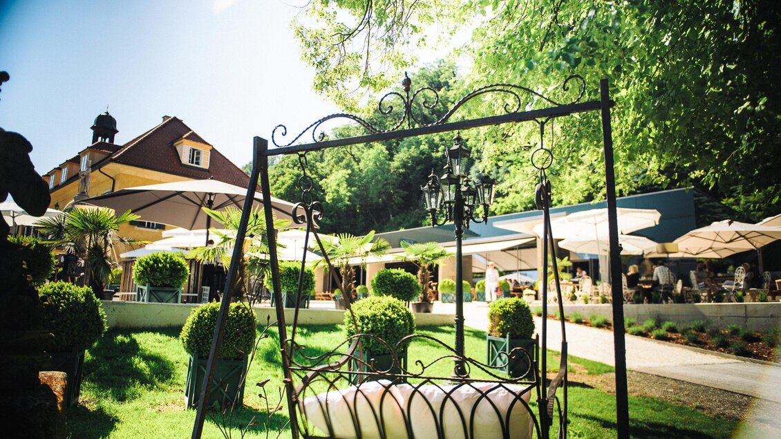 Ein schöner Garten mit einer Schaukel und gemütlichen Sitzbereichen. Im Hintergrund sind Sonnenschirme und ein Gebäude sichtbar. | © Aiola - Joel Kernasenko