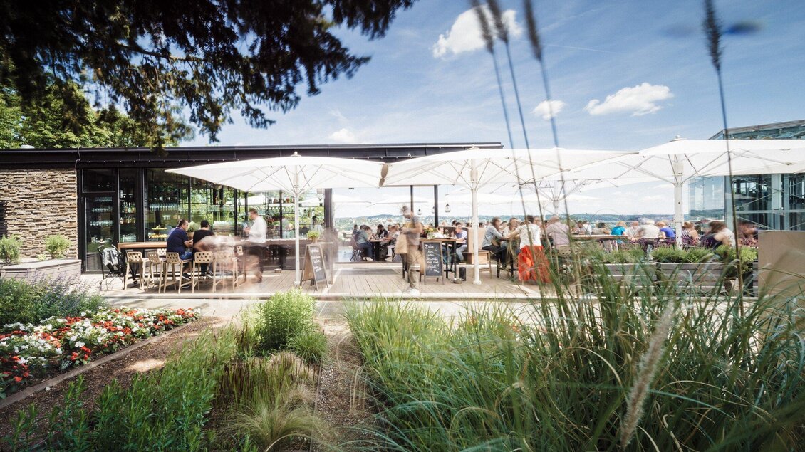Ein modernes Restaurant mit Außenbereich, umgeben von bunten Blumen und Gräsern. Die Gäste genießen die Sonne unter großen Sonnenschirmen. | © aiola