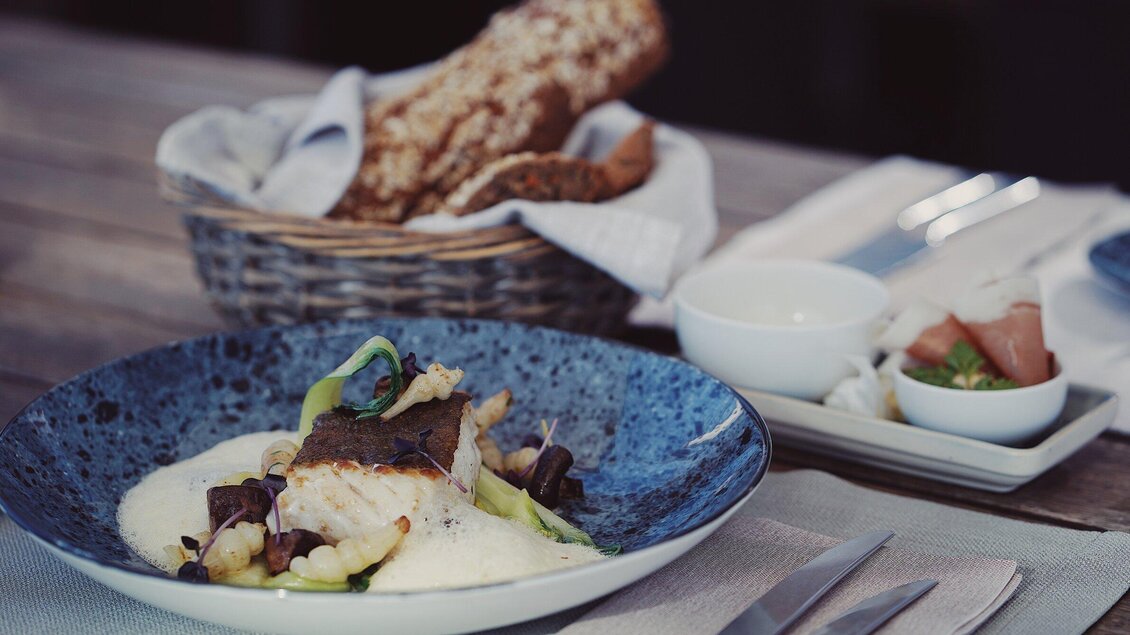 Ein elegantes Gericht mit Fisch, Gemüse und einer cremigen Soße auf einem blauen Teller. Im Hintergrund sind Brot und Beilagen in kleinen Schalen zu sehen. | © aiola