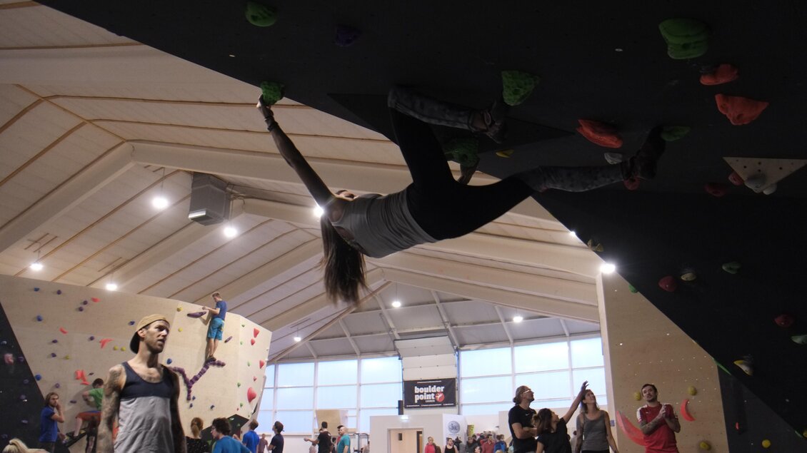 Eine Kletterhalle mit mehreren Personen, die am Bouldern sind. Eine Kletterin ist an der Wand hängend in Aktion zu sehen. | © boulderpoint Leoben