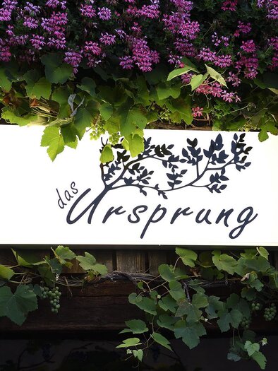 A sign with the inscription "the origin" is surrounded by green ivy and pink flowers. The greenery gives the sign an inviting and natural look. | © Das Ursprung