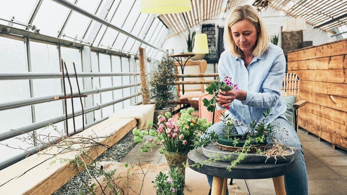 Eine Frau arbeitet in einem Gewächshaus und kümmert sich um Pflanzen. Um sie herum sind verschiedene Blumen und Pflanzen auf einem Tisch angeordnet. | © Armin Walcher