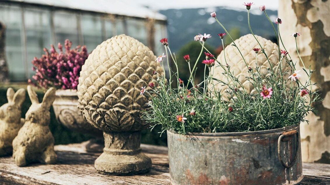 Fliederfarbene Blumen in dekorativen Töpfen auf einem rustikalen Tisch. Im Hintergrund sind zwei kleine Kaninchenfiguren zu sehen. | © Armin Walcher