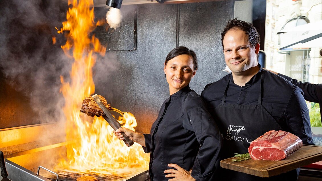 Ein Grill mit großer Flamme und zwei Köchen in der Vordergrund. Die Köchin hält ein Stück Fleisch, während der Koch lächelt und frisches Fleisch präsentiert. | © El Gaucho - Werner Krug