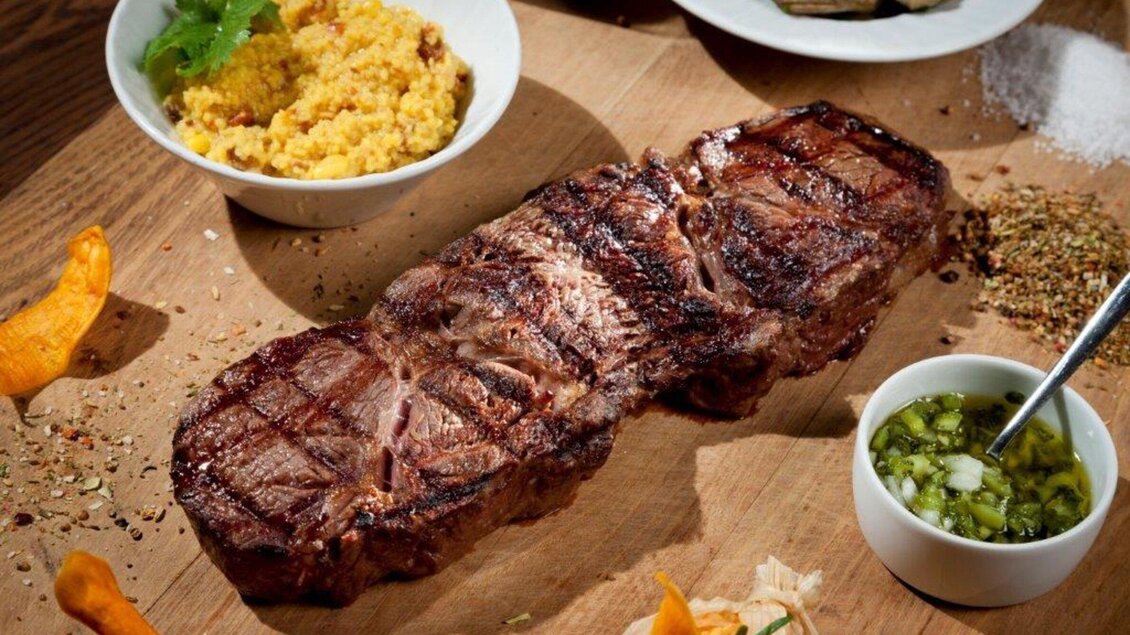 Ein saftiges Steak, perfekt gegrillt, liegt auf einem Holzbrett. Daneben befinden sich Beilagen wie Couscous und Saucen. | © El Gaucho